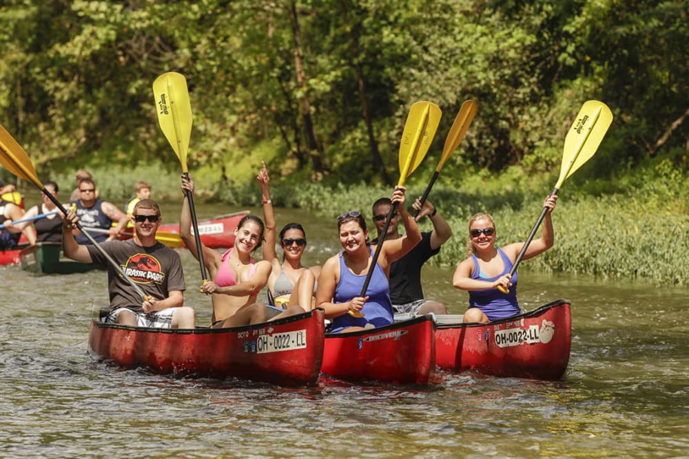A group of six people in canoes paddles while smiling and waving in a scenic river setting.