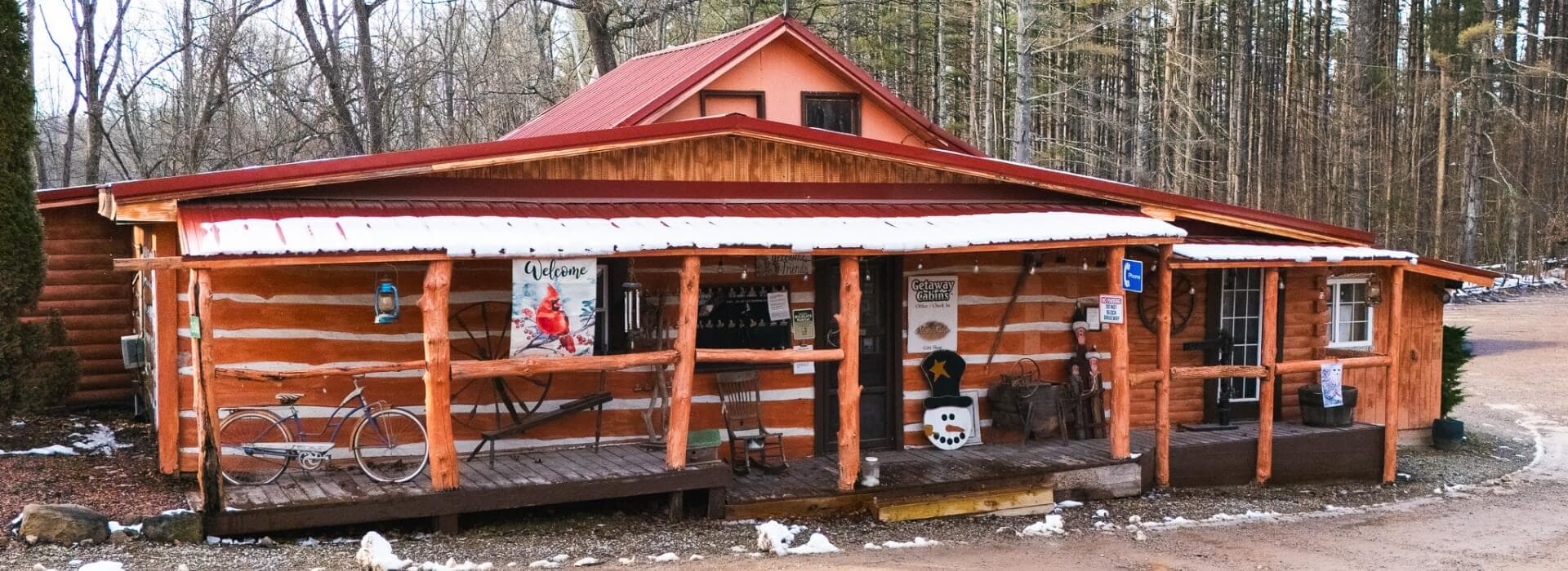 A rustic log cabin with a red metal roof, featuring a porch and a bike outside, surrounded by trees.