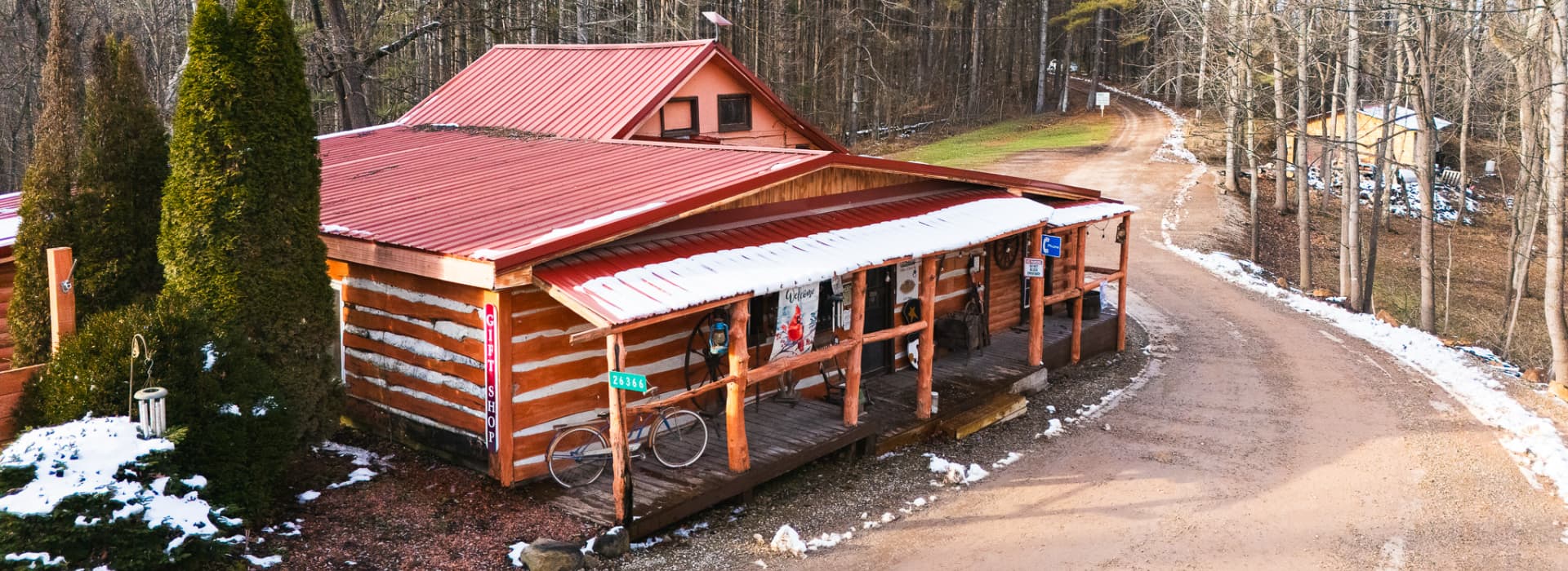 A log cabin with a red metal roof sits along a winding dirt road, surrounded by trees and patches of snow.