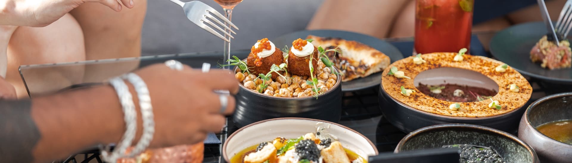 A table spread with various gourmet dishes and drinks, as hands engage with the food.