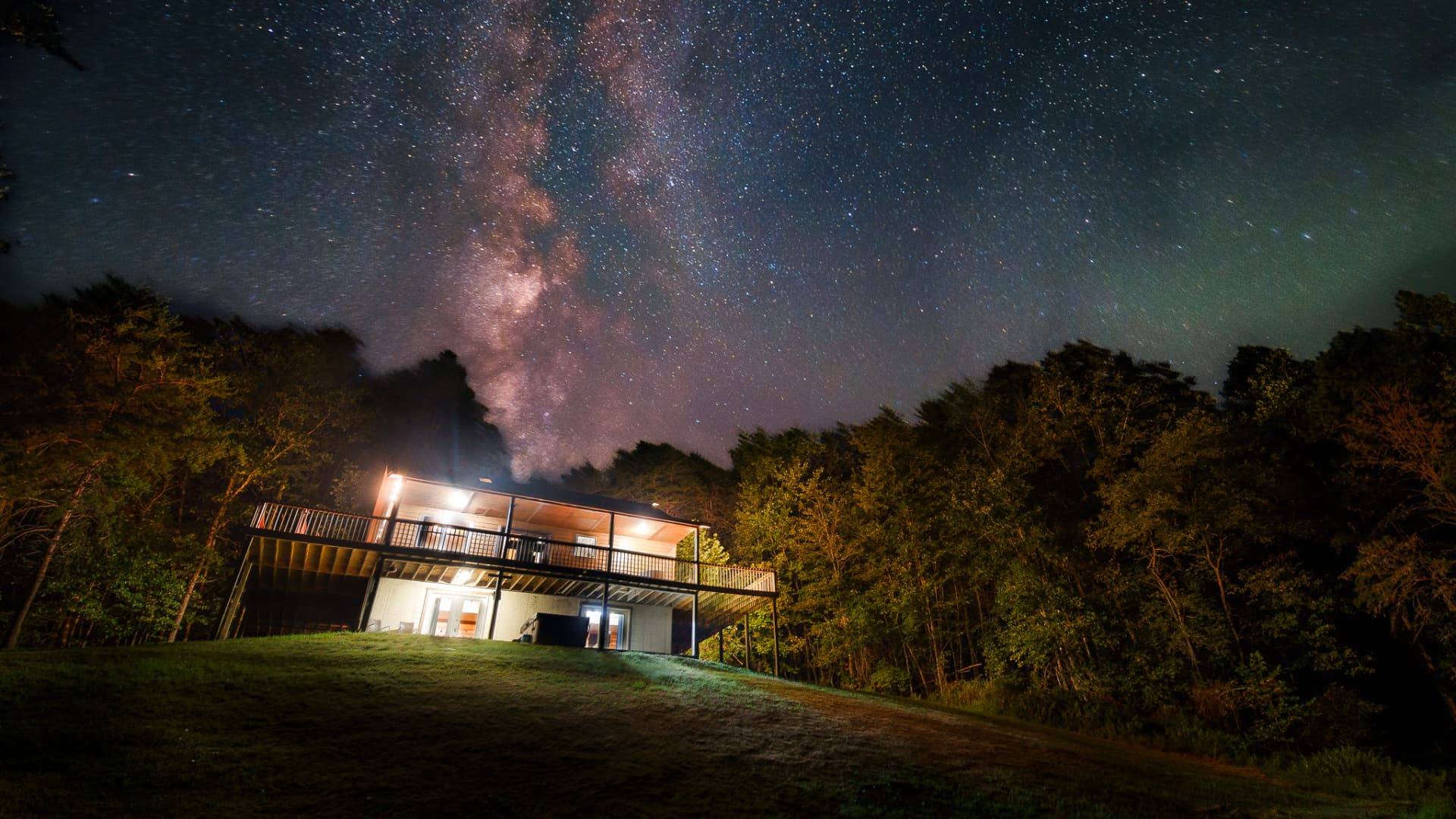 A modern house sits on a grassy hillside under a star-filled sky, featuring the Milky Way.